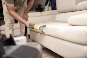 Person cleaning a light-colored sofa with a handheld vacuum cleaner for stain removal in a living room.