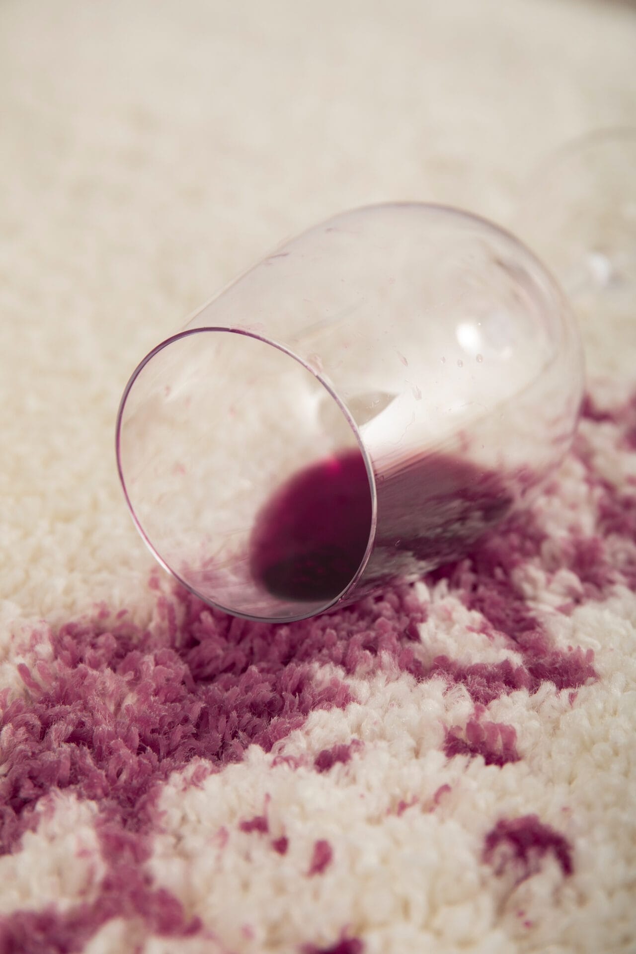 A tipped wine glass spills red wine onto a white carpet, leaving a noticeable rug stain that calls for immediate stain removal.