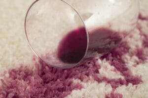 A glass of red wine tipped over on a white carpet, causing a dark red stain—perfect for Fiber Care Solutions' professional cleaning and repair services.