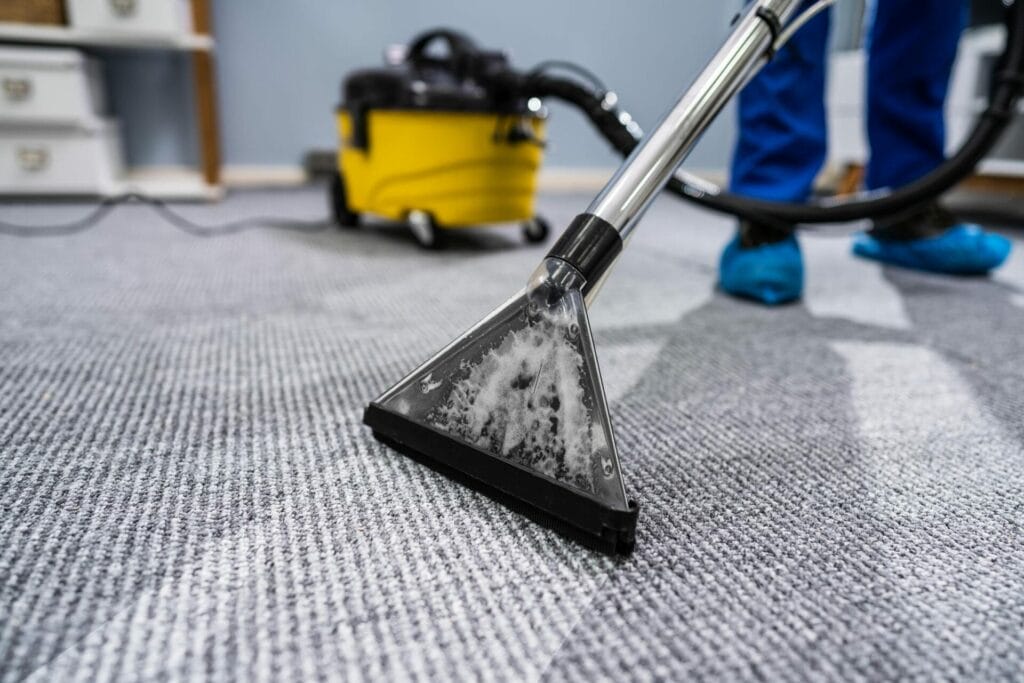 A person cleaning a grey carpet with a vacuum steam cleaner; yellow vacuum visible in the background, highlighting professional carpet cleaning.